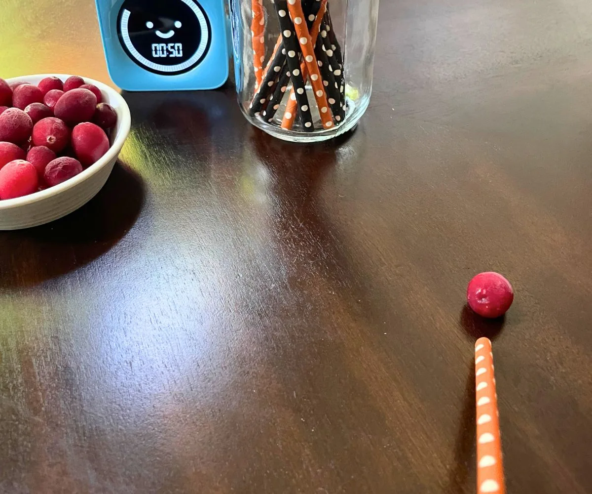 person holding orange straw blowing a cranberry across the table with 50 seconds left on timer