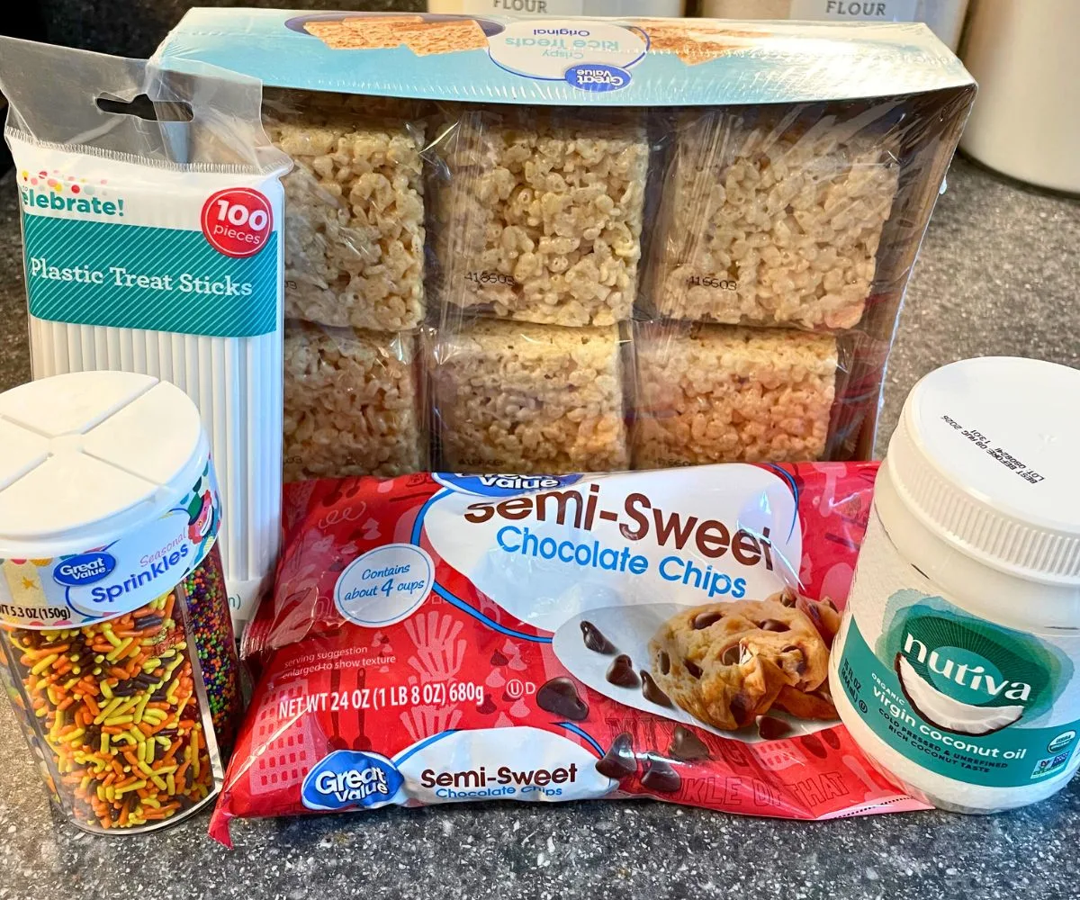 premade Rice Krispies Treats box, plastic treat sticks, semi-sweet chocolate chips, coconut oil, and sprinkles on counter