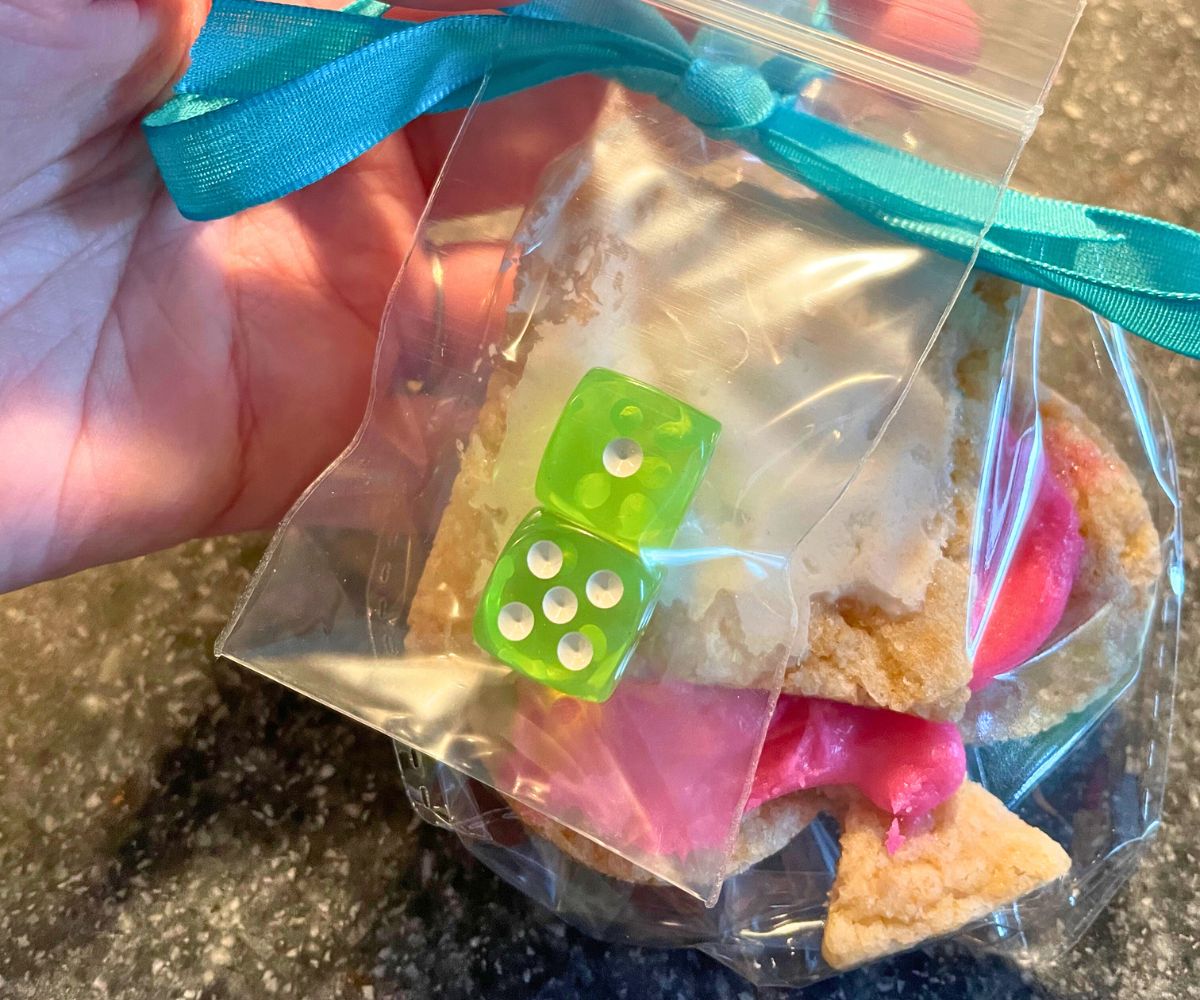 hand holding two playing card suit cookies in bag with two neon green dice on outside, blue ribbon around