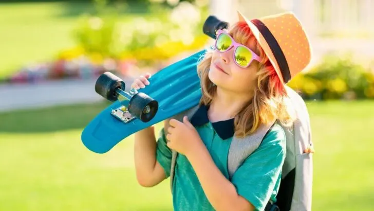 boy in brightly colored clothes smiling looking up at sky, holding skateboard