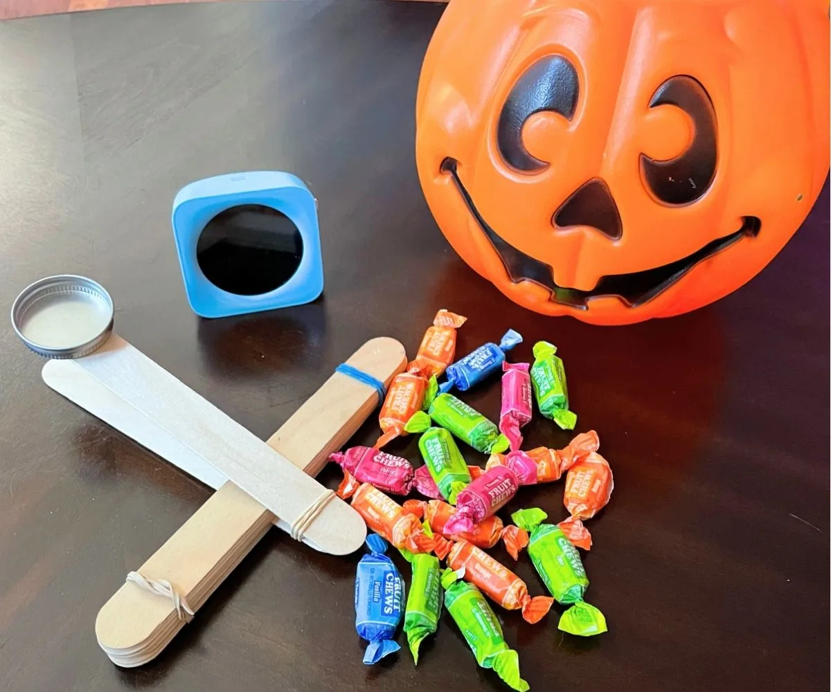 orange pumpkin candy pail, candies, homemade catapult, and timer on dark table