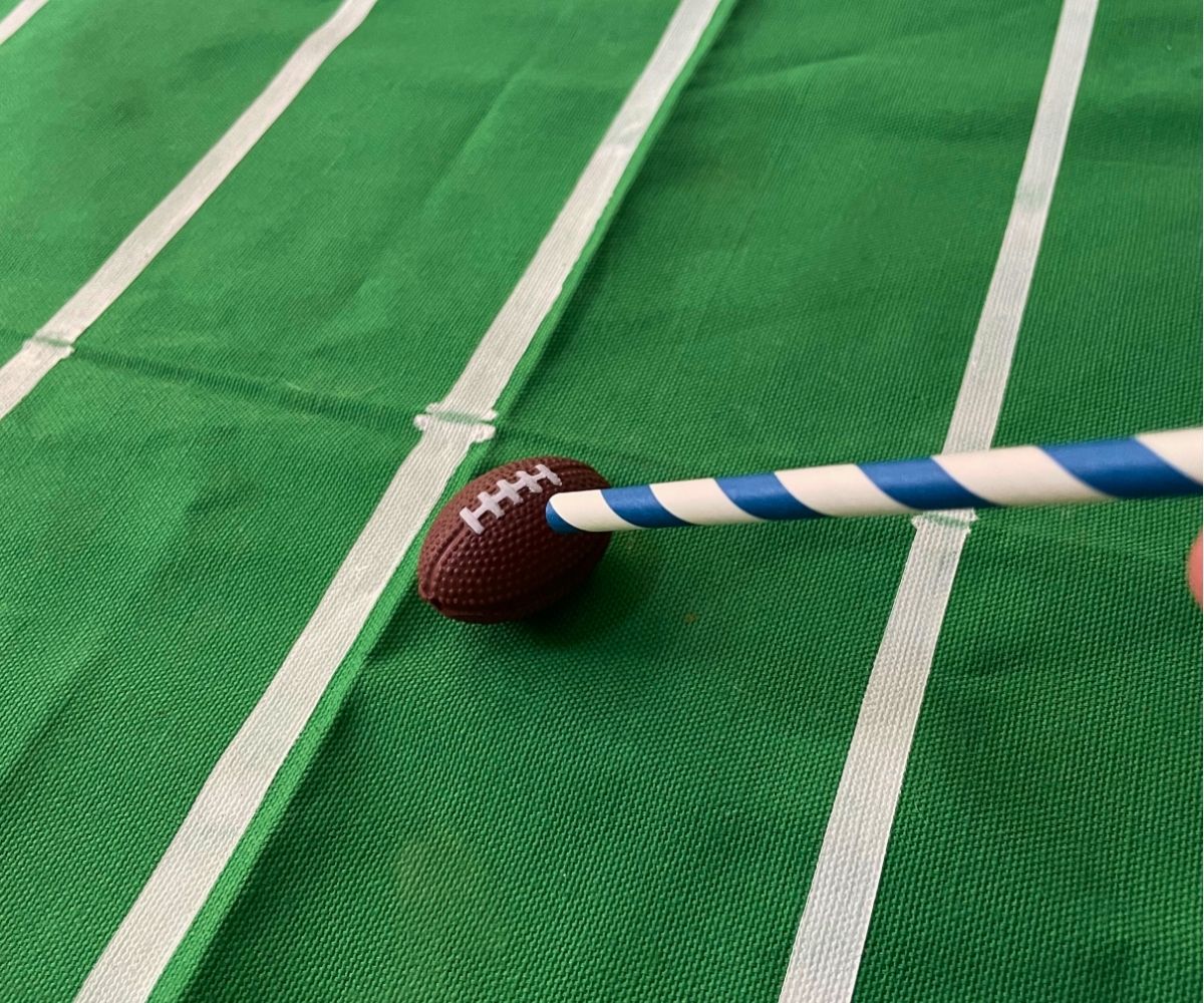 person holding colorful straw to blow into it and move the toy football across the "field"