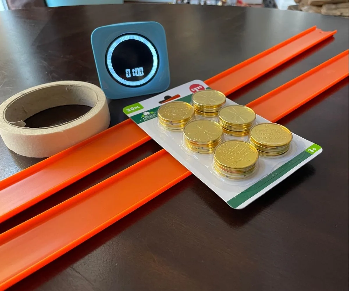 package of gold coins, masking tape, two bright orange race track segments, and a timer set at 1 minute