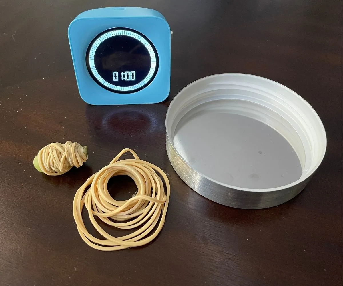 little figurine wrapped in lots of rubber bands, next to rubber bands, container lid, and timer