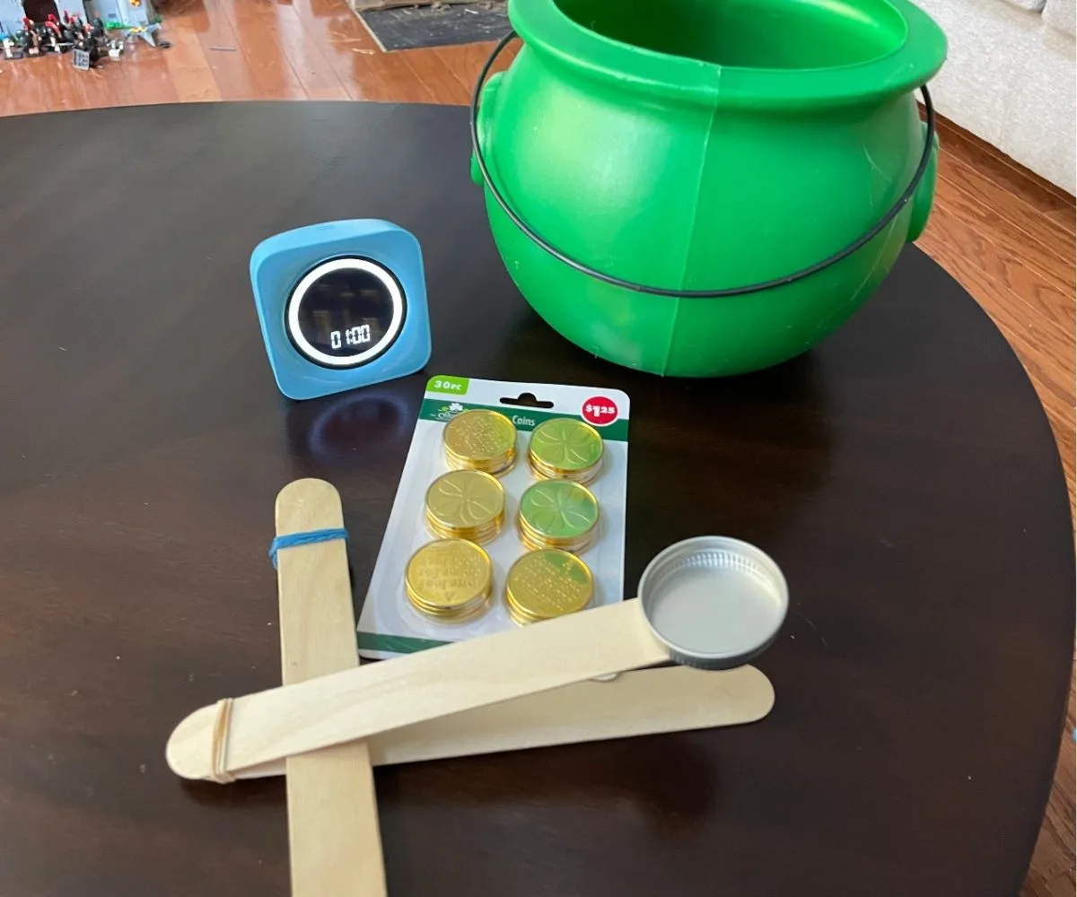 gold coins, timer for 1 minute, homemade catapult and green plastic leprechaun pot on table