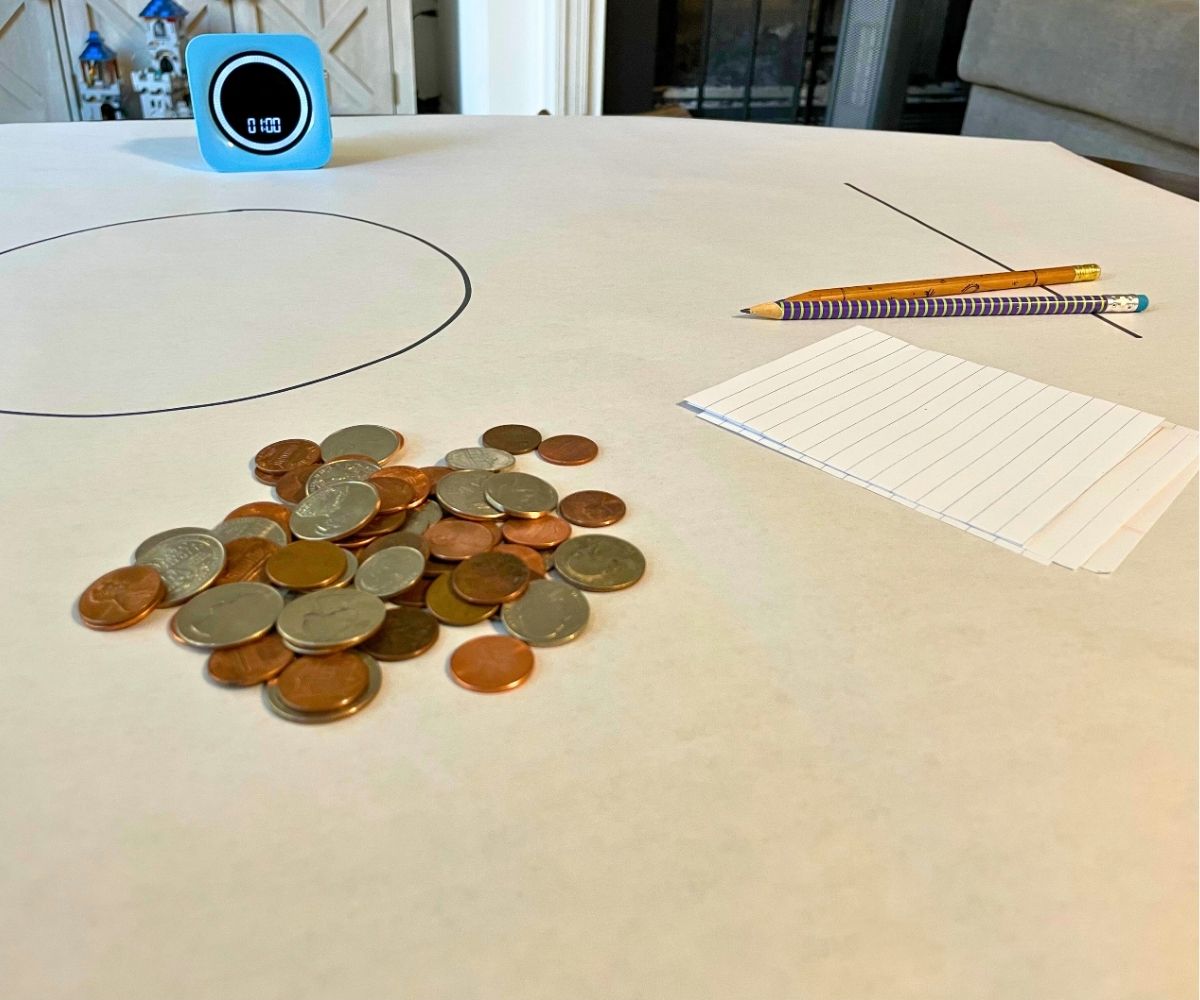 big white paper with big circle in middle, timer, two pencils, scratch paper, and big pile of coins