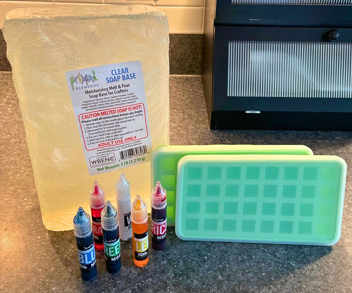 clear soap base, 6 tubs of soap dye, and two small cube molds on kitchen counter