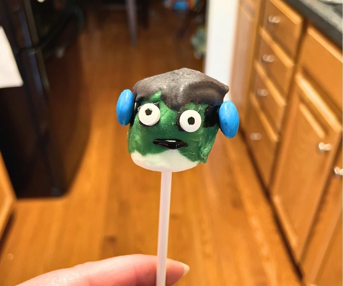 Green and black Frankenstein marshmallow on stick with two blue M&Ms on either side of head