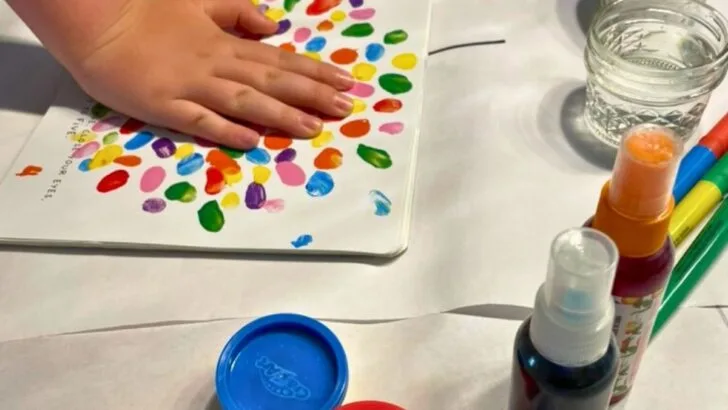kid hand pushing down on colorful book page, with lots of color play things around him