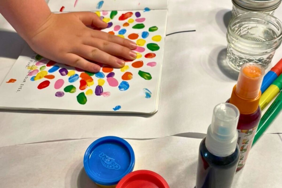 kid hand pushing down on colorful book page, with lots of color play things around him