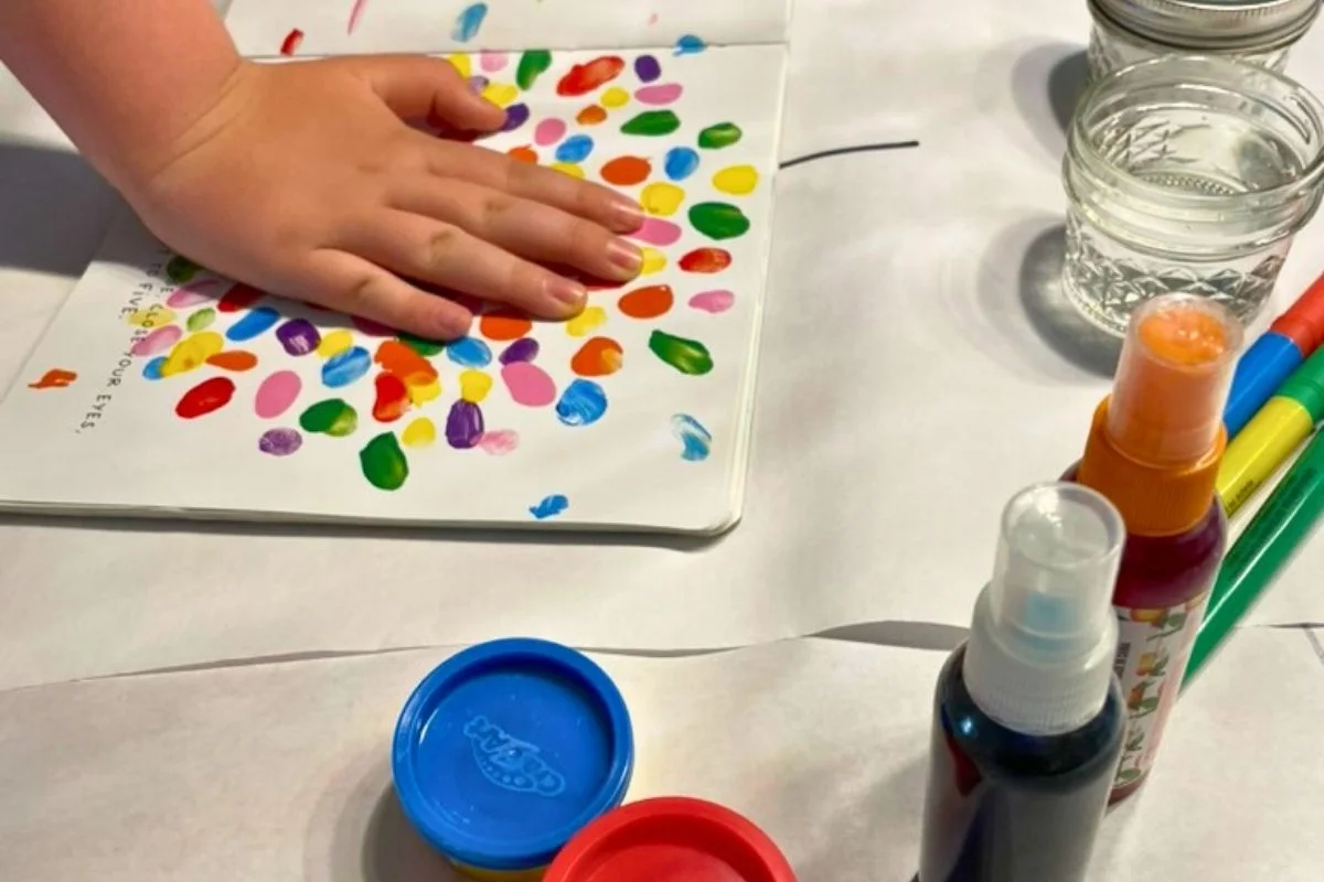 kid hand pushing down on colorful book page, with lots of color play things around him