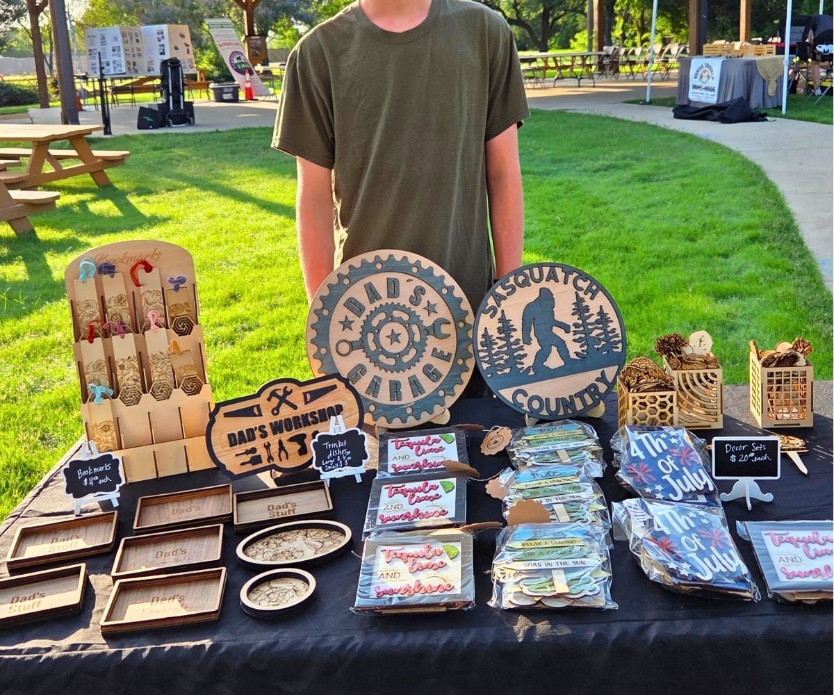 Asher standing behind his table of lots of CO2 laser printing products at a farmer's market