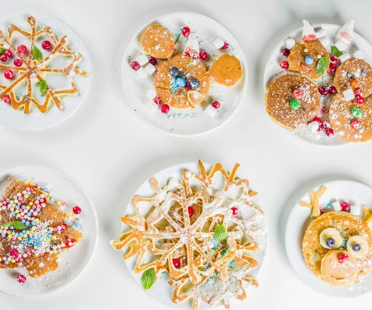 six different white plates filled with fun, holiday-shaped pancakes, pieces of candy, and powdered sugar