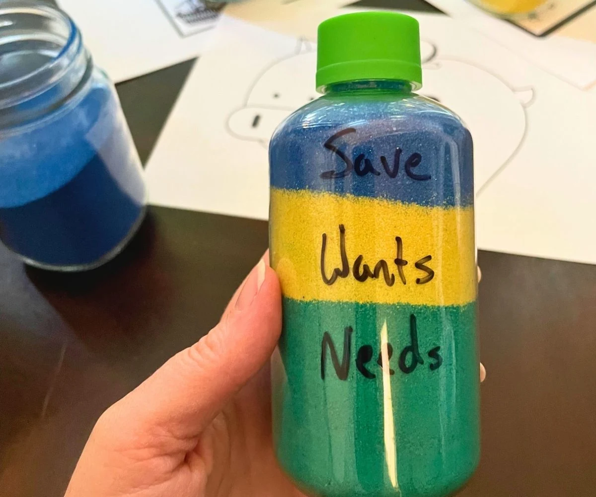 hand holding a three-colored sand jar with "Needs", "Wants", and "Save" written on different layers