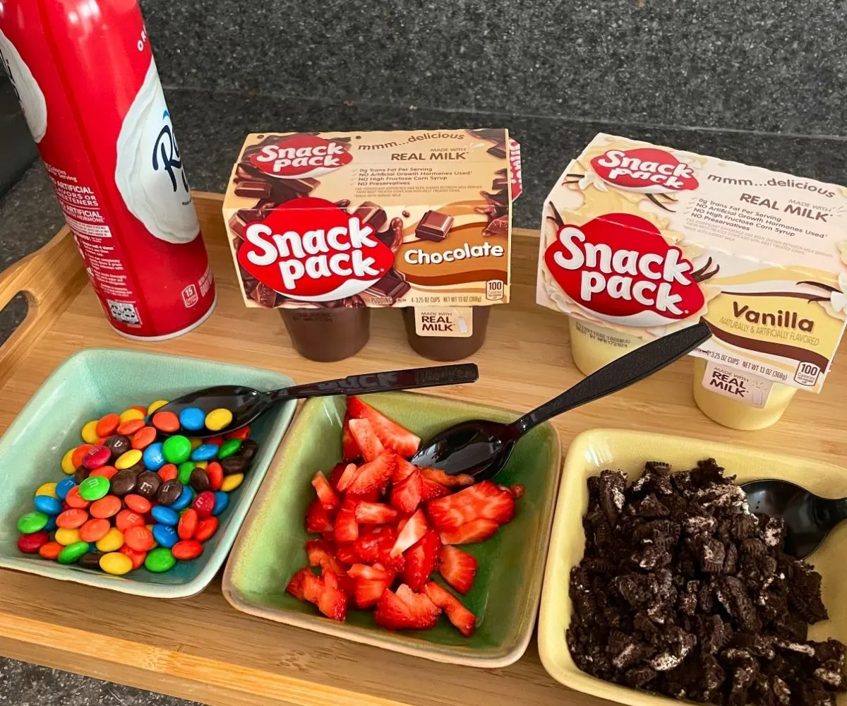 pudding snack packs, whipped cream, and three toppings on light wooden tray