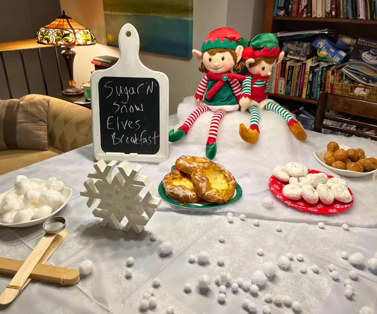 white tablecloth on table with large elves sitting up on cotton batting, lots of treats and cotton snowballs
