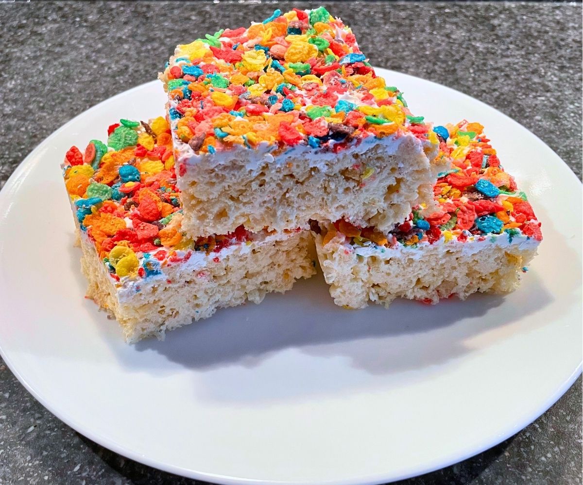 three rice krispies treats on white plate with layer of white candy melt and Fruity Pebbles on top