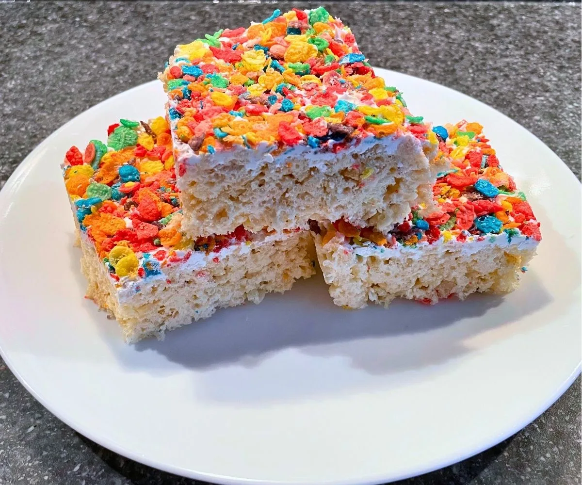 three rice krispies treats on white plate with layer of white candy melt and Fruity Pebbles on top