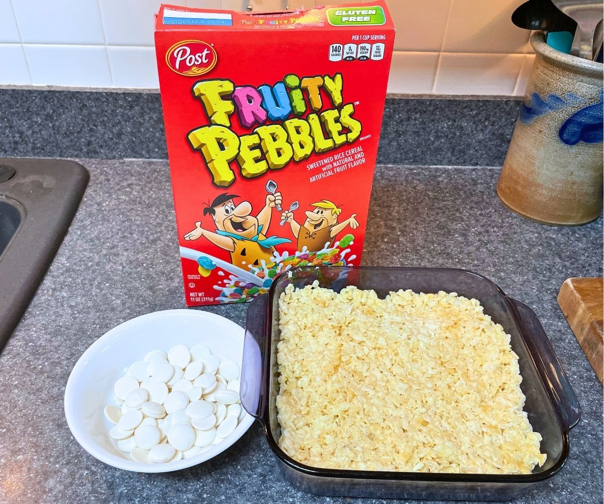 Baked Rice Krispies treats in pan with white candy melts next to it and a box of Fruity Pebbles
