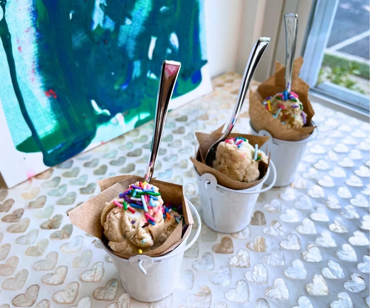 three white little buckets lined with parchment paper filled with mounds of rainbow-sprinkled edible cookie dough with silver spoons