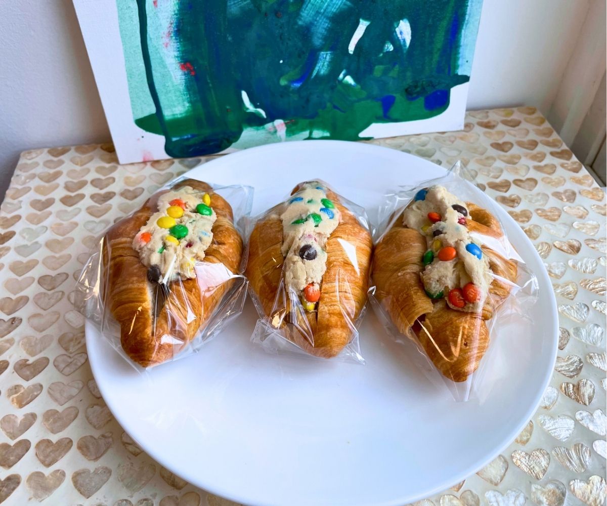 three croissants filled with M&M cookie dough and wrapped in plastic on white plate to sell