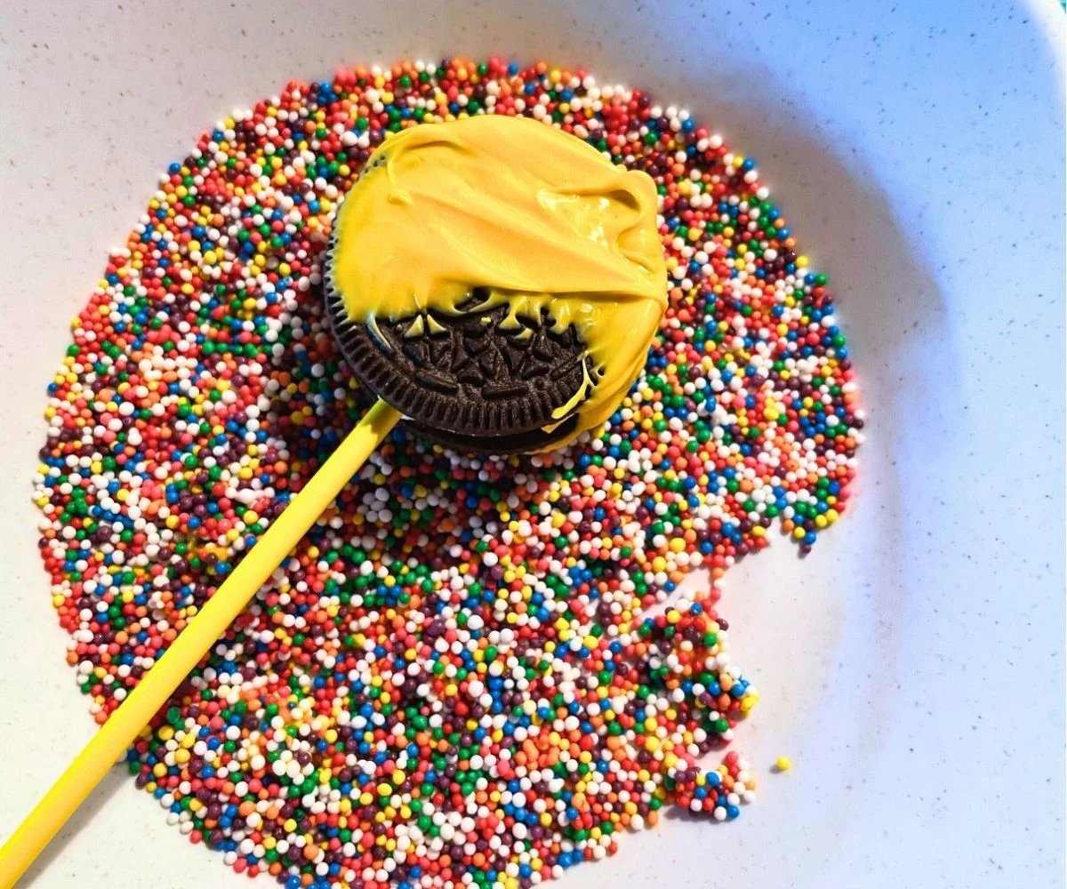 yellow candy-melt-covered Oreo on yellow lollipop stick in rainbow covered sprinkles dish