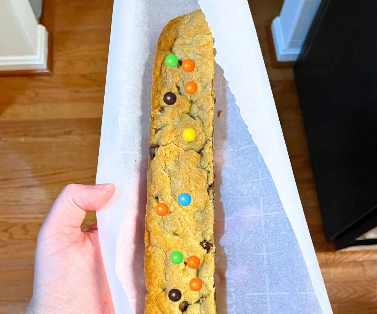 someone holding a footlong, rainbow M&M cookie bar in parchment paper