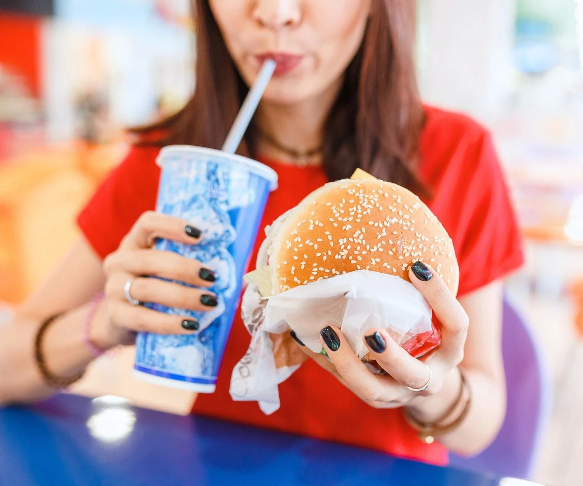 teen girl with black manicured nails eating big cheeseburger and large drink
