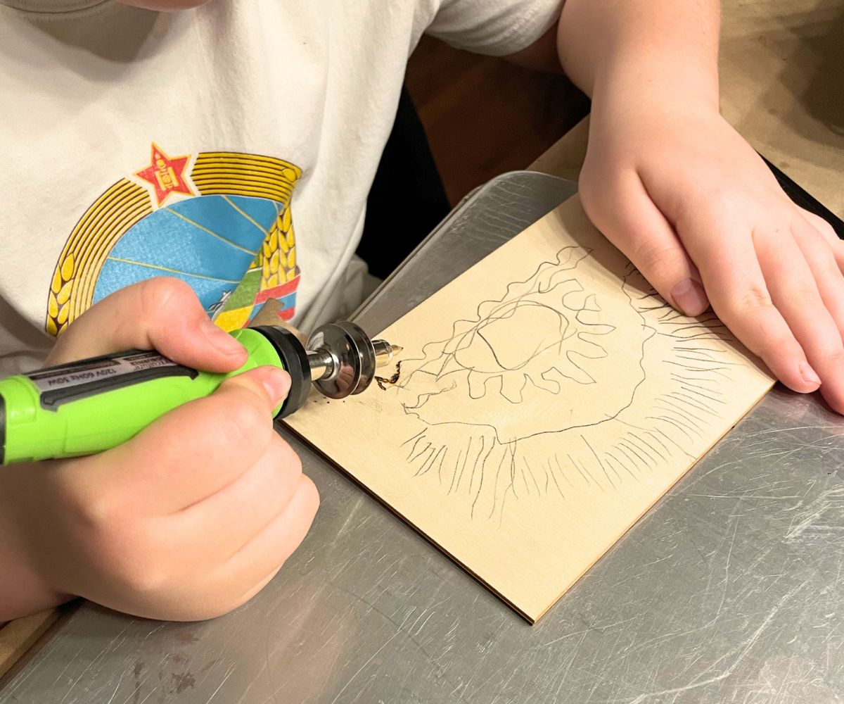 10 year old boy holding a wood burning tool and burning a design on wood on top of a sheet pan