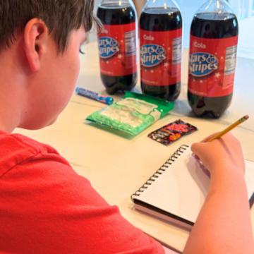 child making prediction in notebook in front of 3 2-liters of off-brand cokes and some candies
