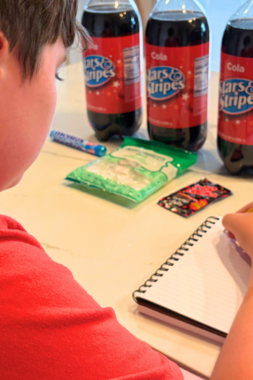 child making prediction in notebook in front of 3 2-liters of off-brand cokes and some candies