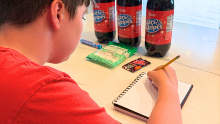 child making prediction in notebook in front of 3 2-liters of off-brand cokes and some candies