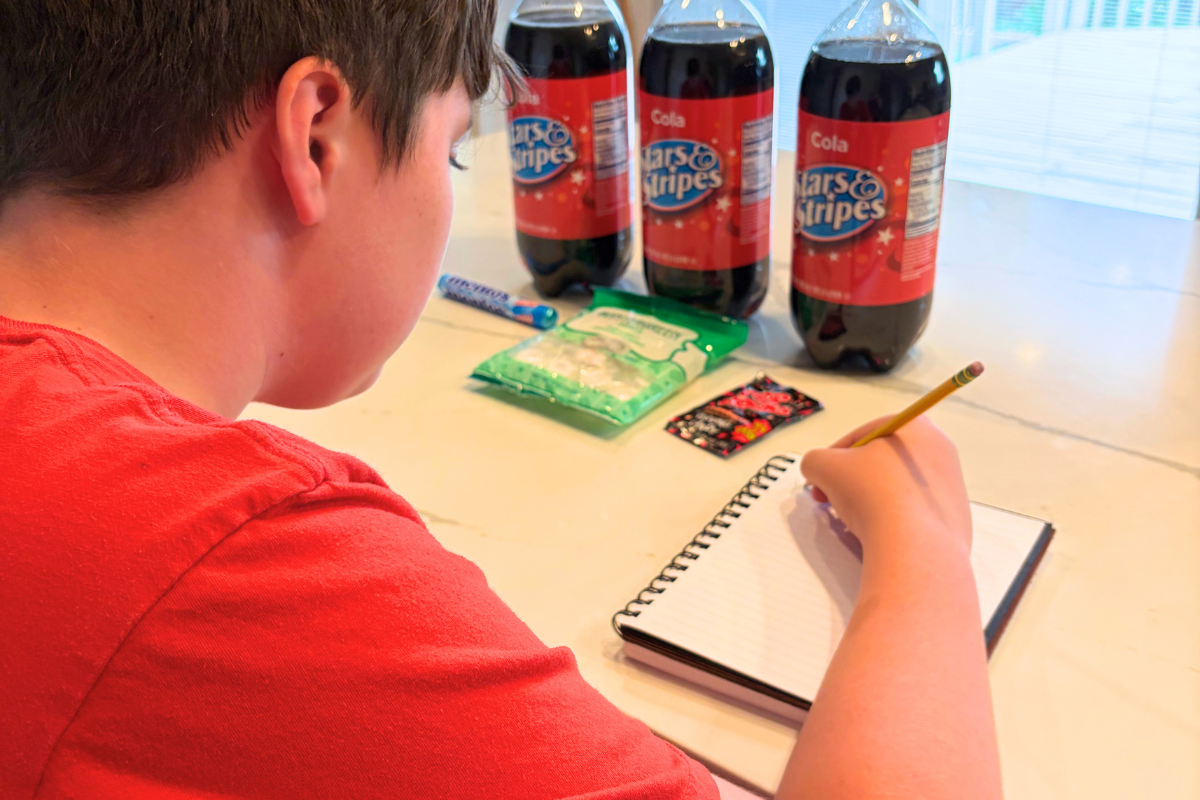 child making prediction in notebook in front of 3 2-liters of off-brand cokes and some candies
